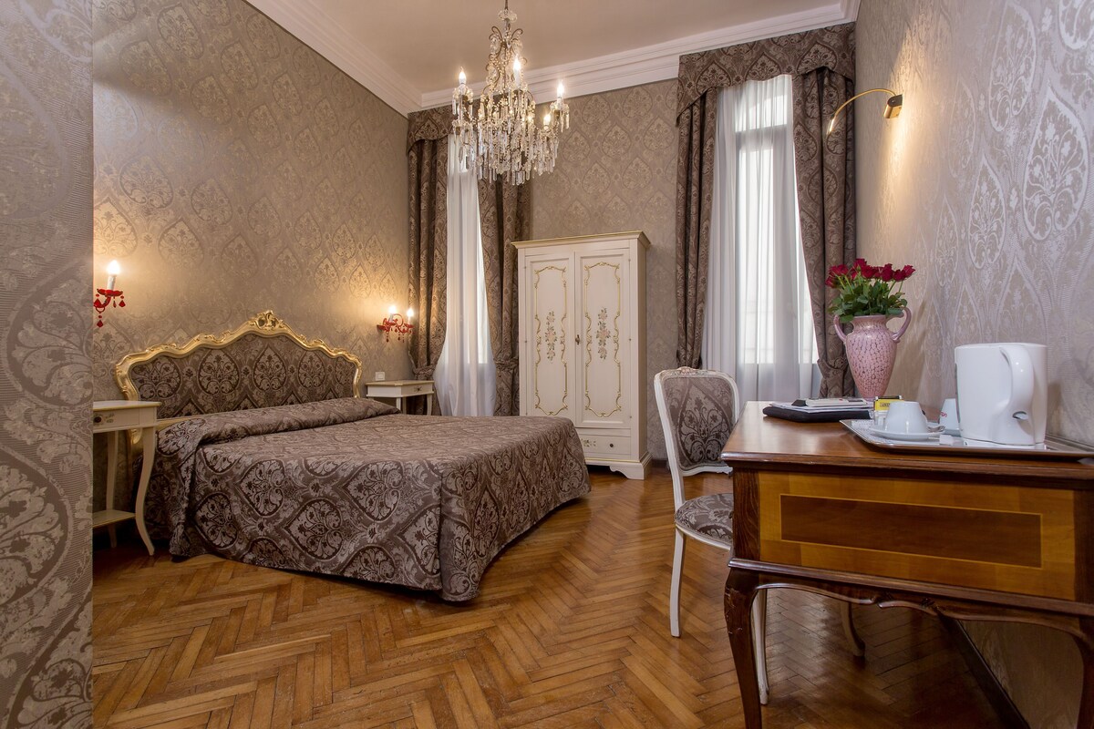 Habitación de hotel con cama, un escritorio con silla, armario, candelabro y un jarrón con flores.