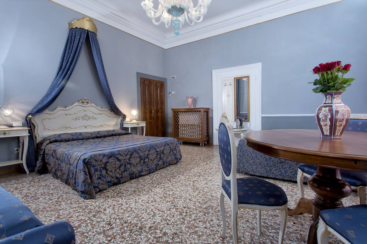A bedroom with a canopy bed, a round wooden table, a vase with flowers, and a mirror on the wall.