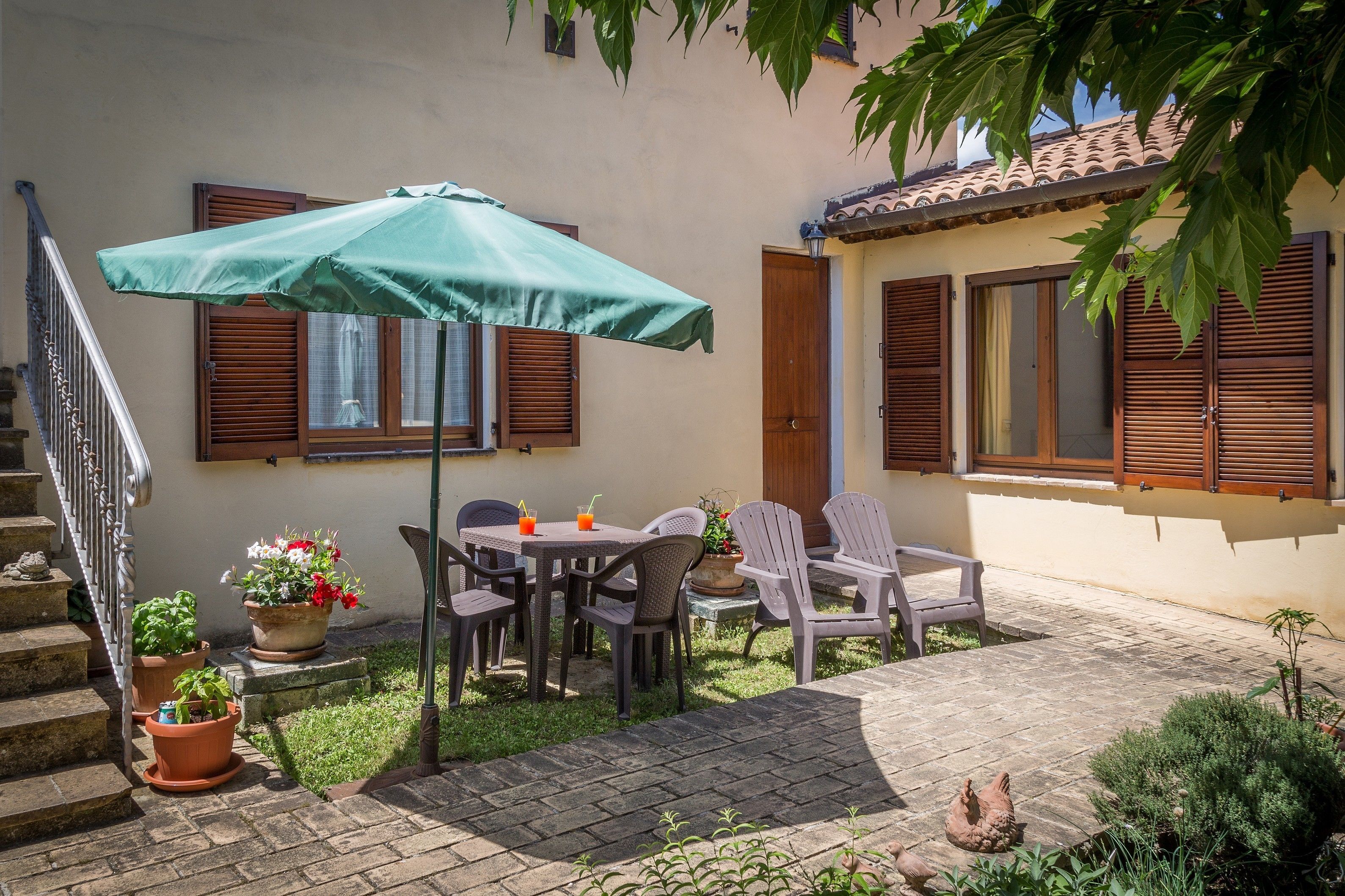 Un patio con una mesa, sillas, un parasol verde, plantas en macetas y una escalera.