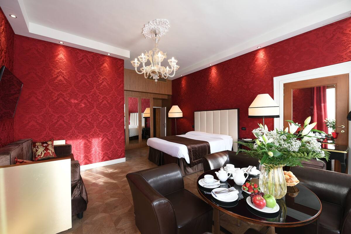 Quarto de hotel com parede estampada em vermelho, uma cama, área de estar com mesa posta para o café da manhã e um lustre.