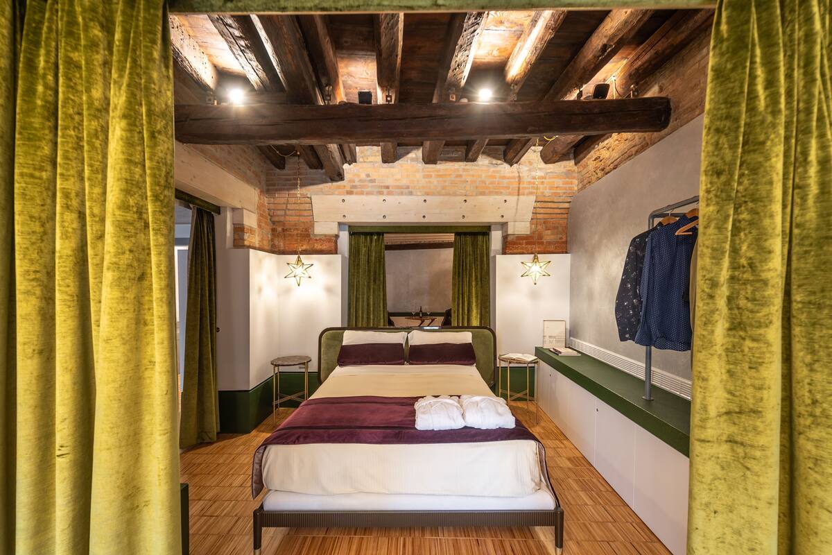 Quarto com teto de madeira, parede de tijolos, cama com cortinas verdes e um porta-roupas.