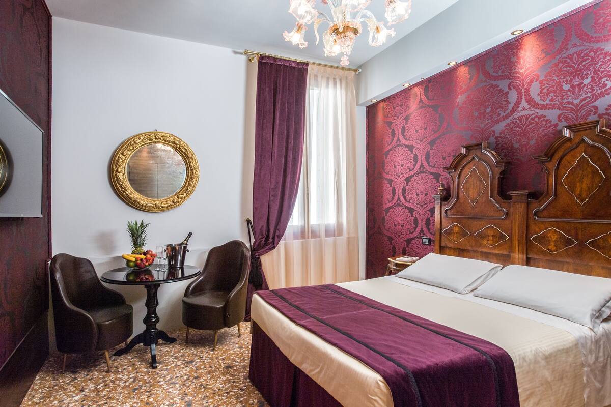 Quarto de hotel com uma cama grande, duas cadeiras, uma mesinha com frutas e uma garrafa, e um espelho na parede.
