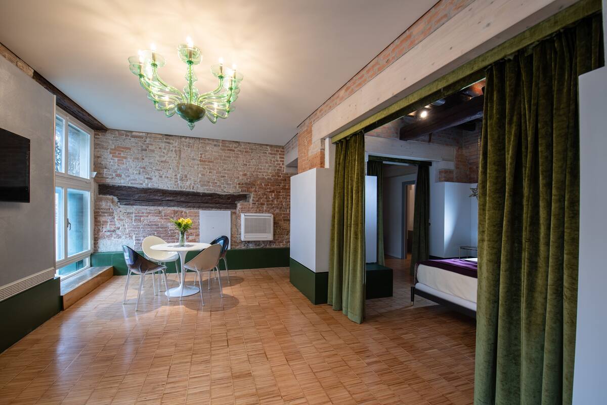 Quarto com parede de tijolos, piso de madeira, mesa de jantar com cadeiras, lustre e cama com cortinas verdes.