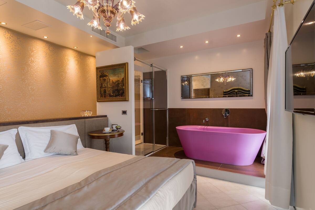 Quarto de hotel moderno com cama grande, chuveiro amplo, banheiro com paredes de vidro e um lustre.