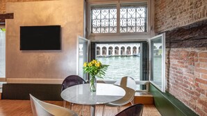 A dining area with a round table, chairs, and a vase of flowers. A large window with a wrought iron gate provides a view of a canal and buildings. - Locanda Leon Bianco on the Grand Canal (Venice)