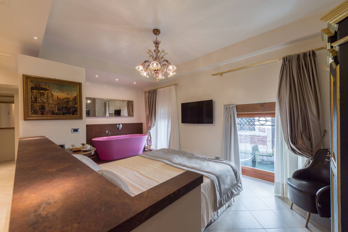 Um quarto moderno com uma cama grande, uma toucador com pia rosa, um candelabro, um quadro na parede e vista pela janela.