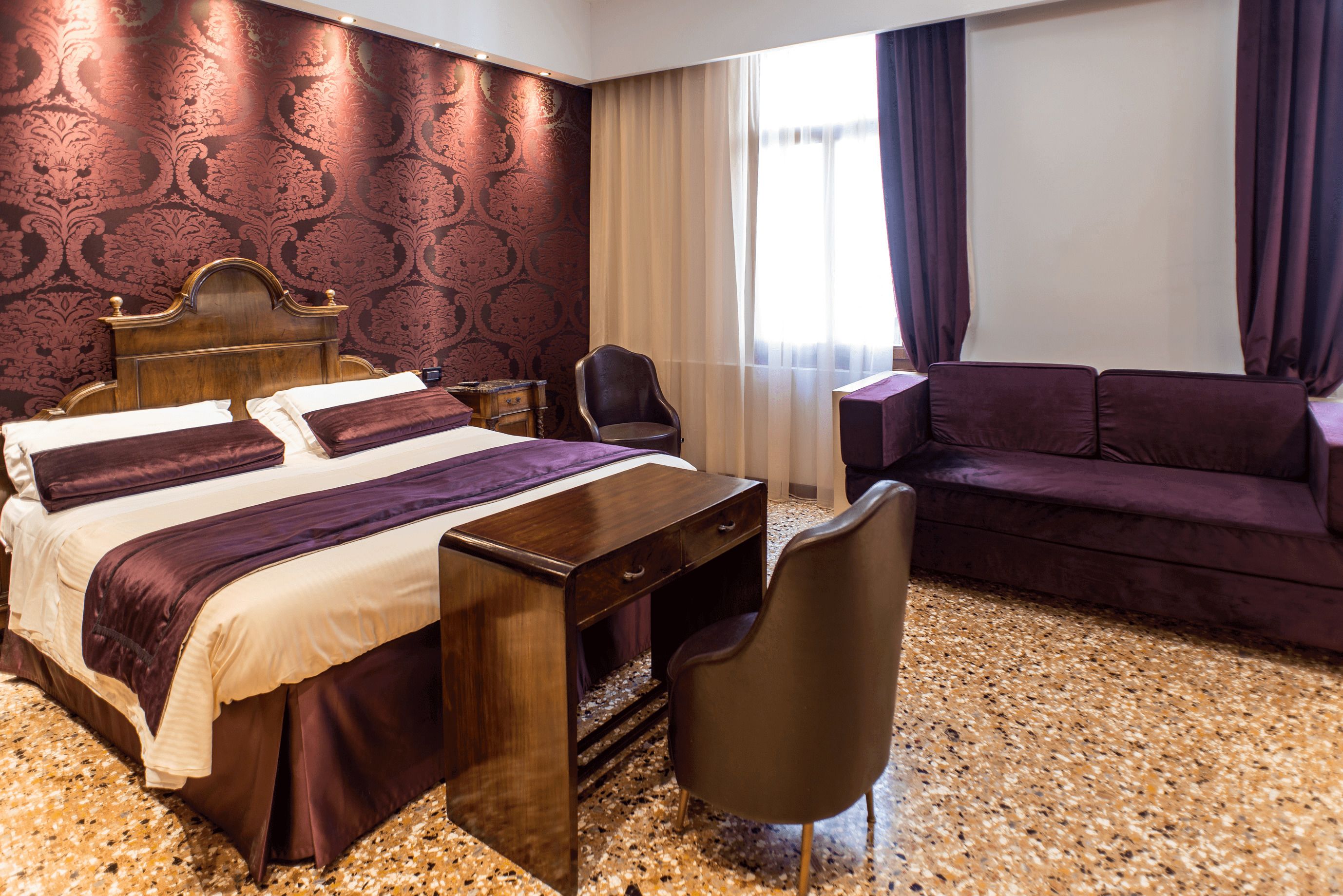 Ein Hotelzimmer mit Bett, Schreibtisch, Sessel, Sofa und einem großen Fenster mit Vorhängen.
