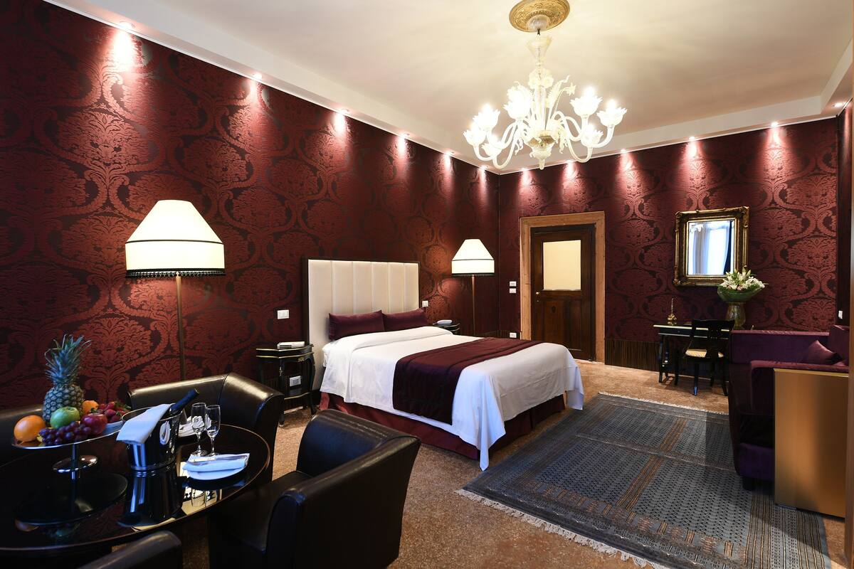 Quarto de hotel com uma cama grande, duas mesas de cabeceira, uma escrivaninha com cadeira, uma mesinha com frutas e uma cafeteira, e um candelabro.