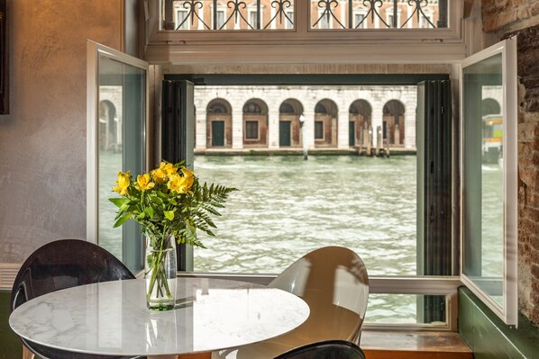 Ein Tisch mit einer Vase gelber Blumen, Blick auf einen Kanal mit Gebäuden und ein Fenster mit einem Metallgeländer.