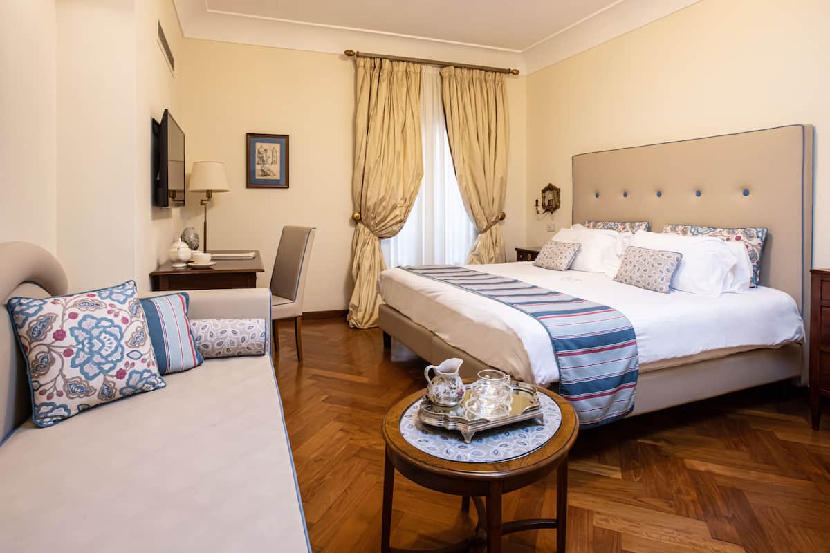 A hotel room with a bed, a sofa, a table with a tray, a lamp, a mirror, and curtains.