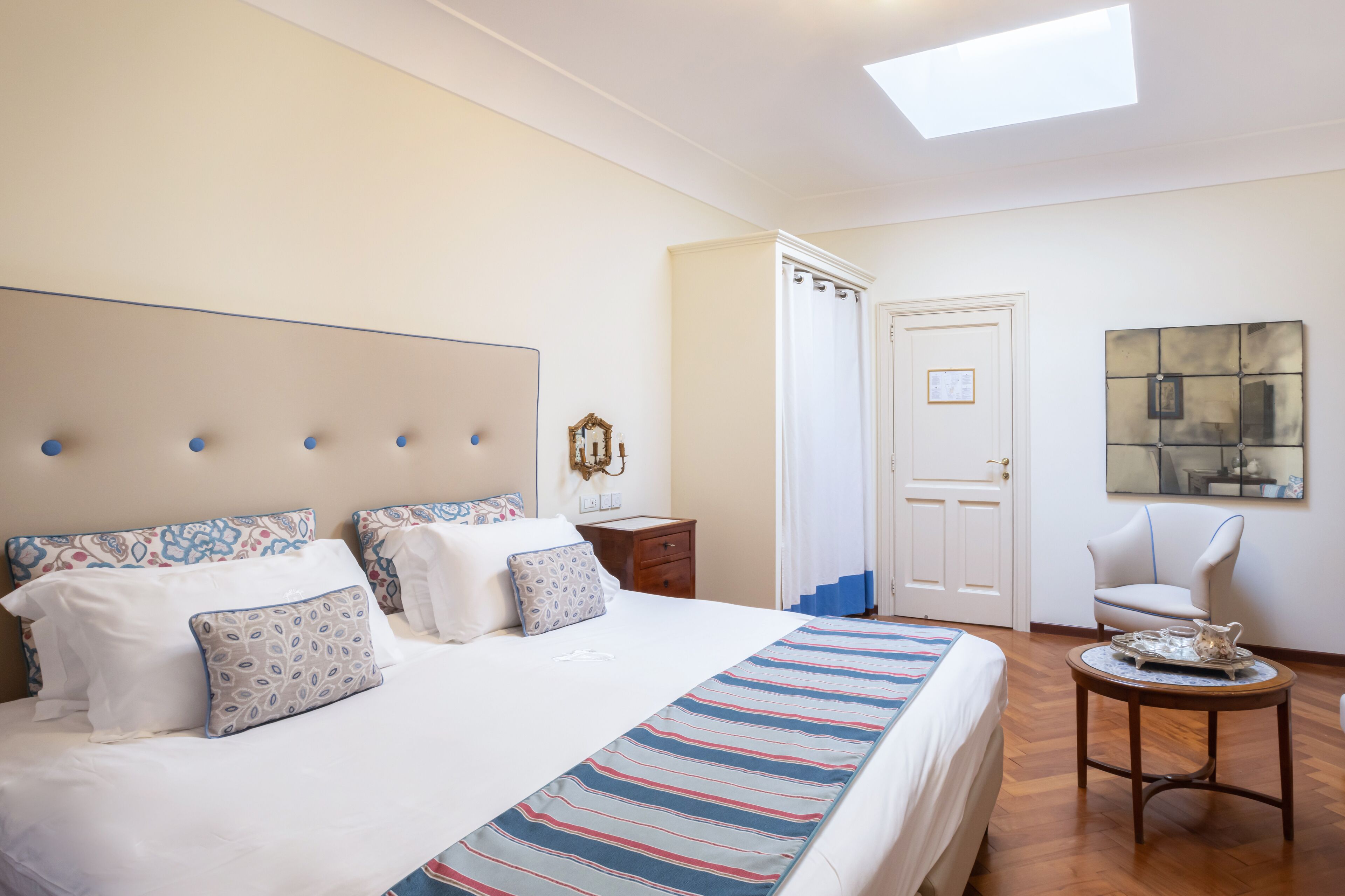 A hotel room with a large bed, a bedside table, a chair, a small table with a tray, and a painting on the wall.