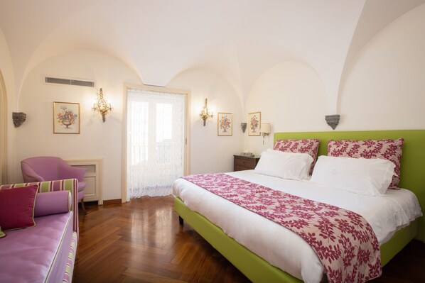 Une chambre avec une tête de lit verte, un grand lit avec une literie à motifs floraux, un fauteuil rose et un plancher en bois.