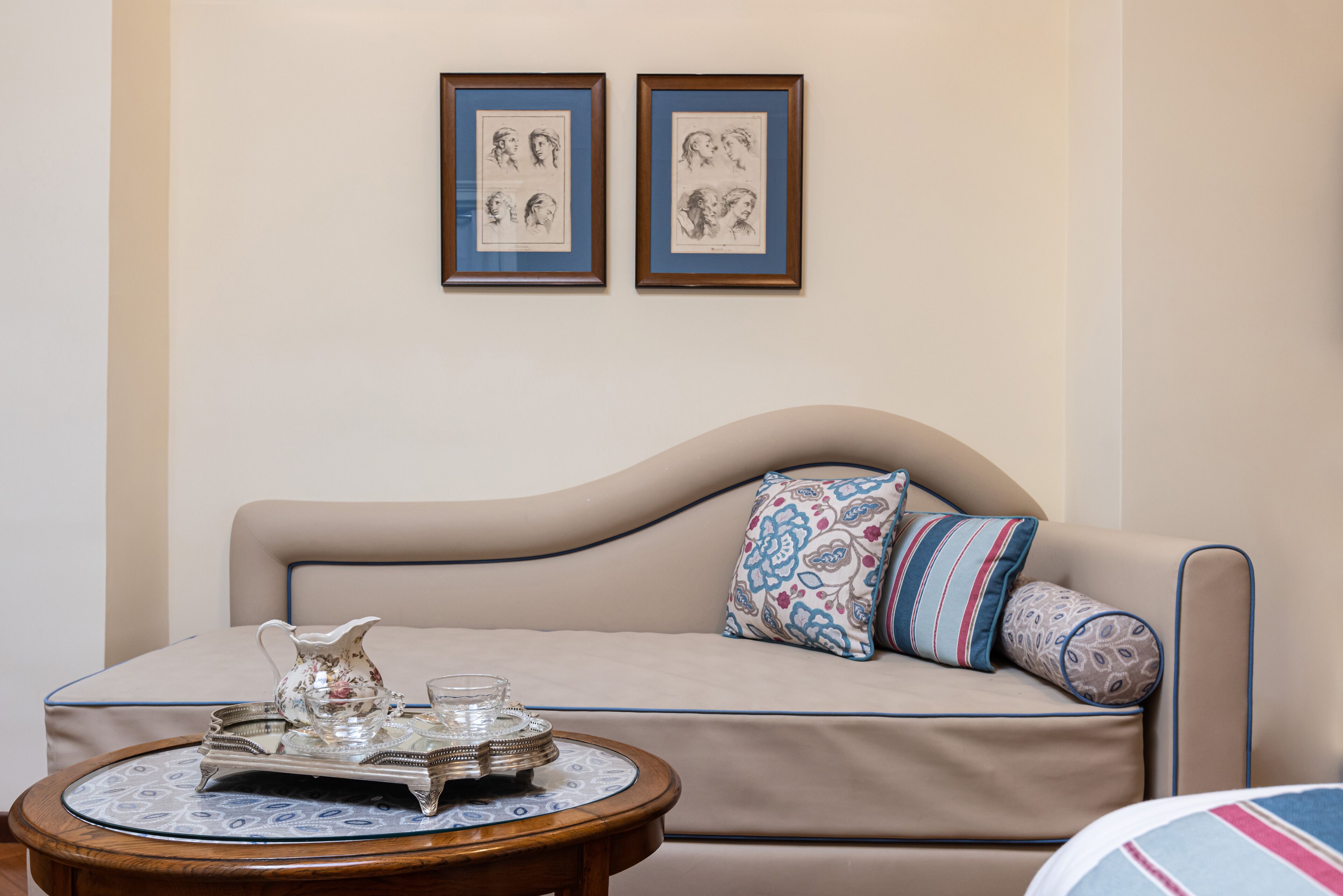 A beige sofa with patterned cushions, a small round table with a tray of silverware and a pitcher, and two framed sketches on the wall.