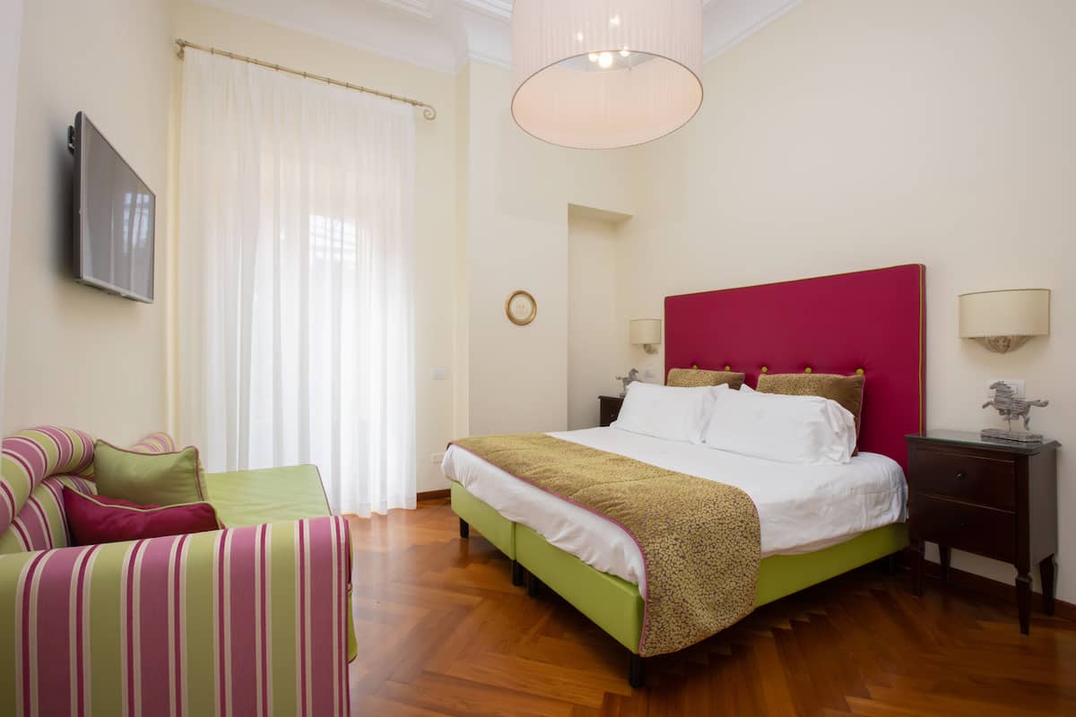 A hotel room with a bed, a green and pink striped sofa, a wooden floor, a large window with curtains, and a flat-screen TV mounted on the wall.