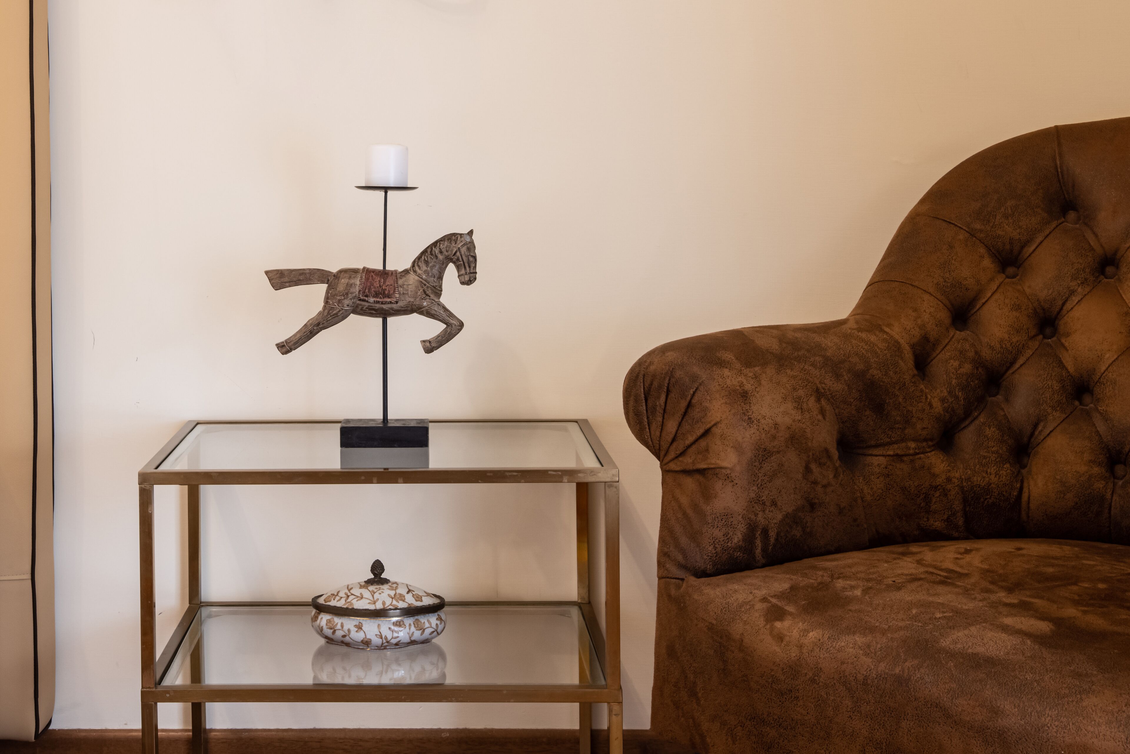 Un fauteuil marron à accoudoirs capitonnés, placé à côté d’une table à plateau en verre, sur laquelle reposent une sculpture équestre décorative et un chandelier.