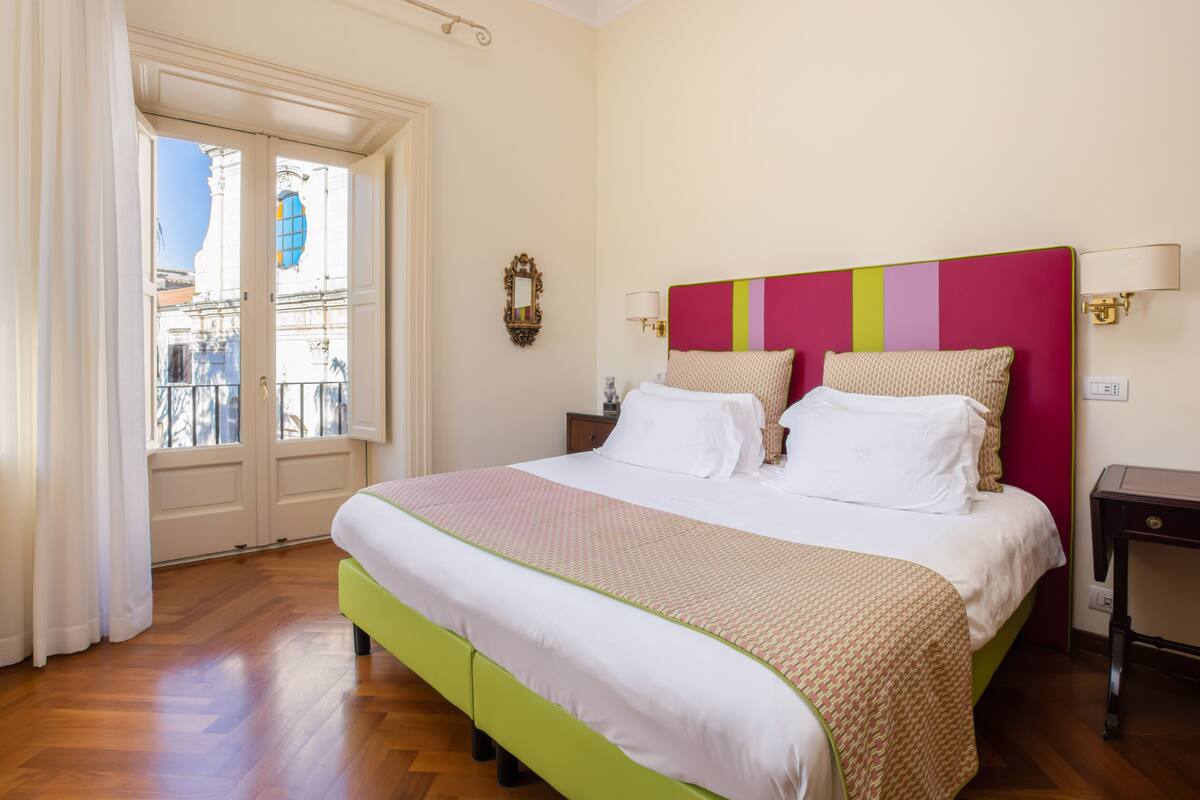 A hotel room with a large bed, a nightstand, a lamp, and a view of the city through a window.