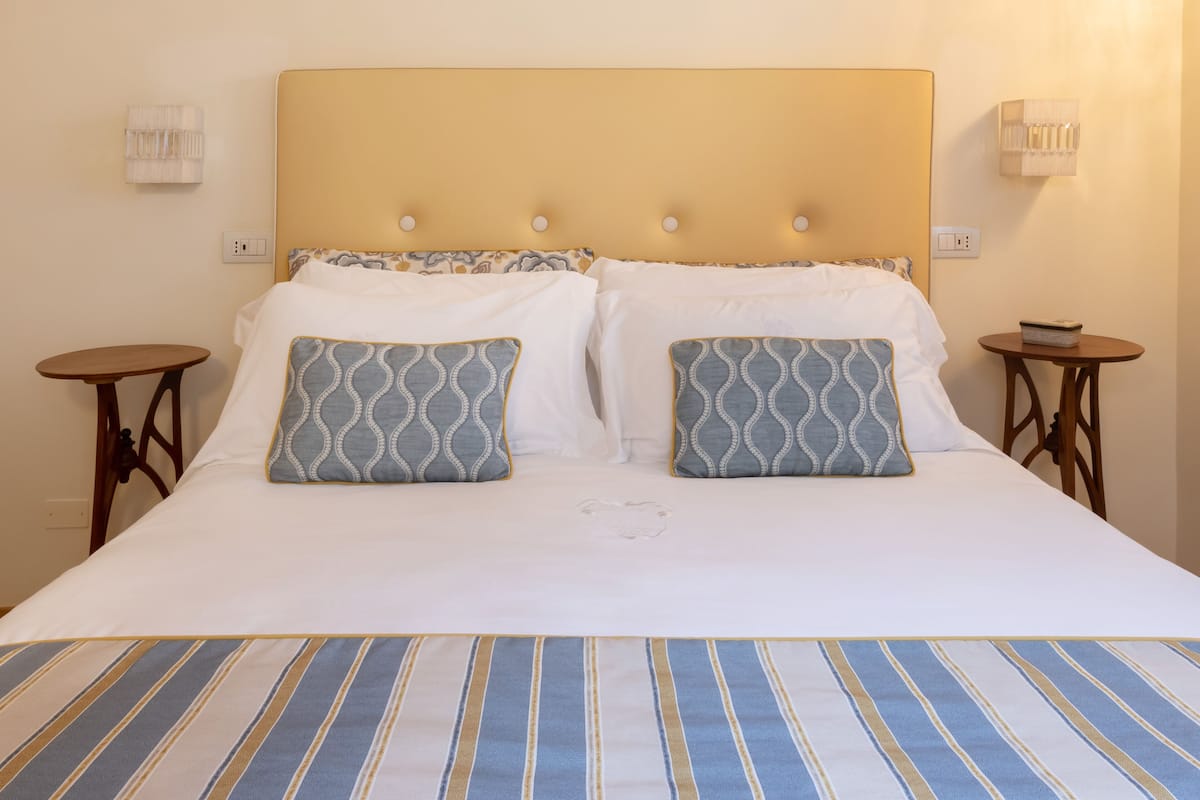 A neatly made bed with a striped bedspread, two patterned pillows, and a headboard with round buttons. There are two small wooden nightstands with lamps on either side of the bed.