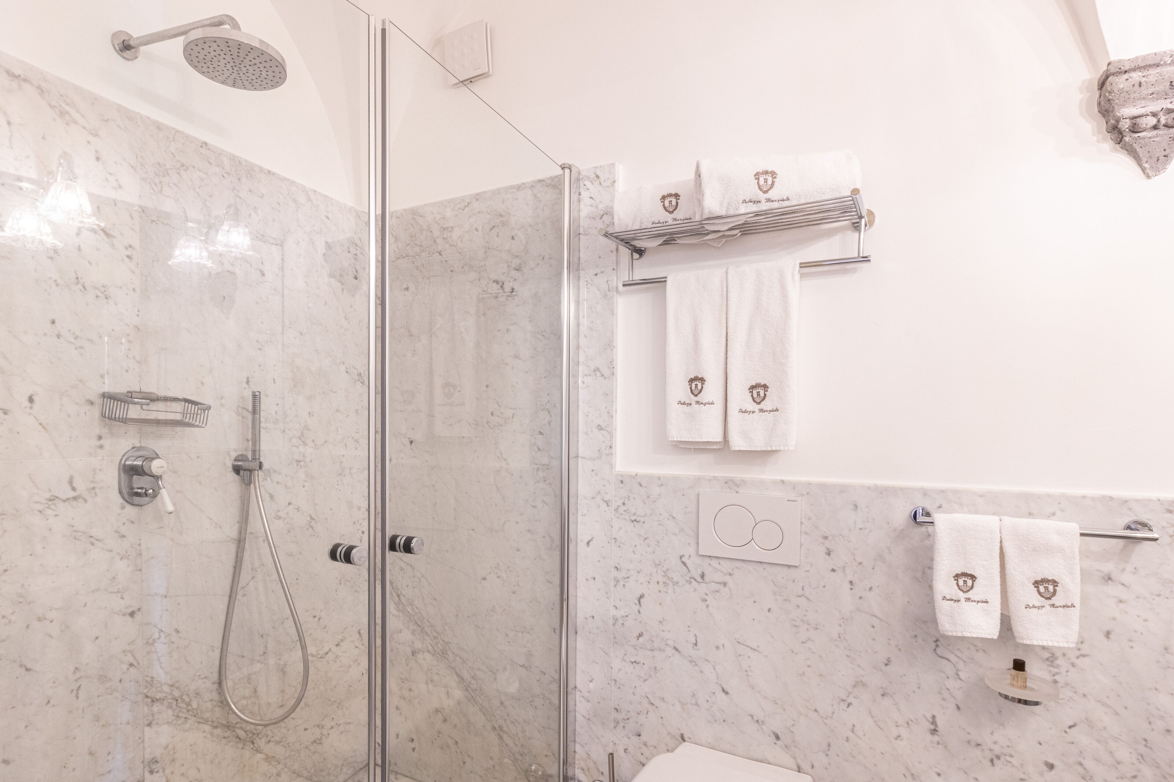 Un baño moderno con ducha envuelta en vidrio, paredes de mármol y toallas con el logotipo de un escudo.