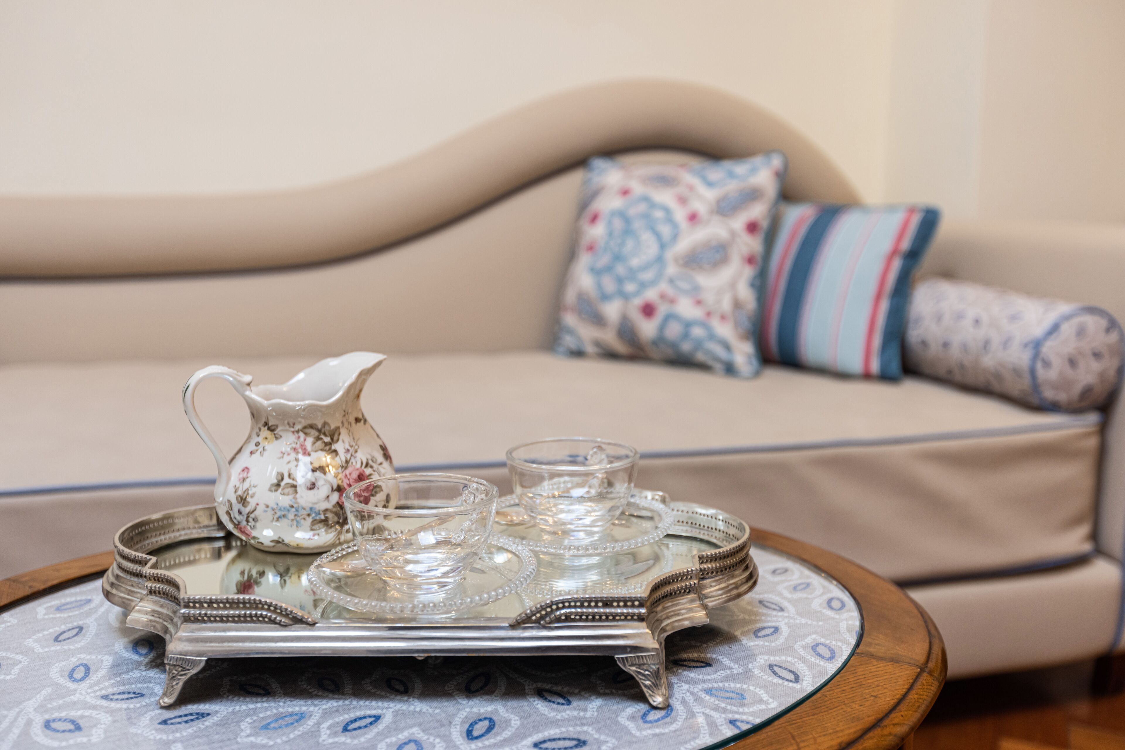A room with a beige sofa, patterned pillows, a tray with a pitcher and glasses, and a patterned tablecloth.