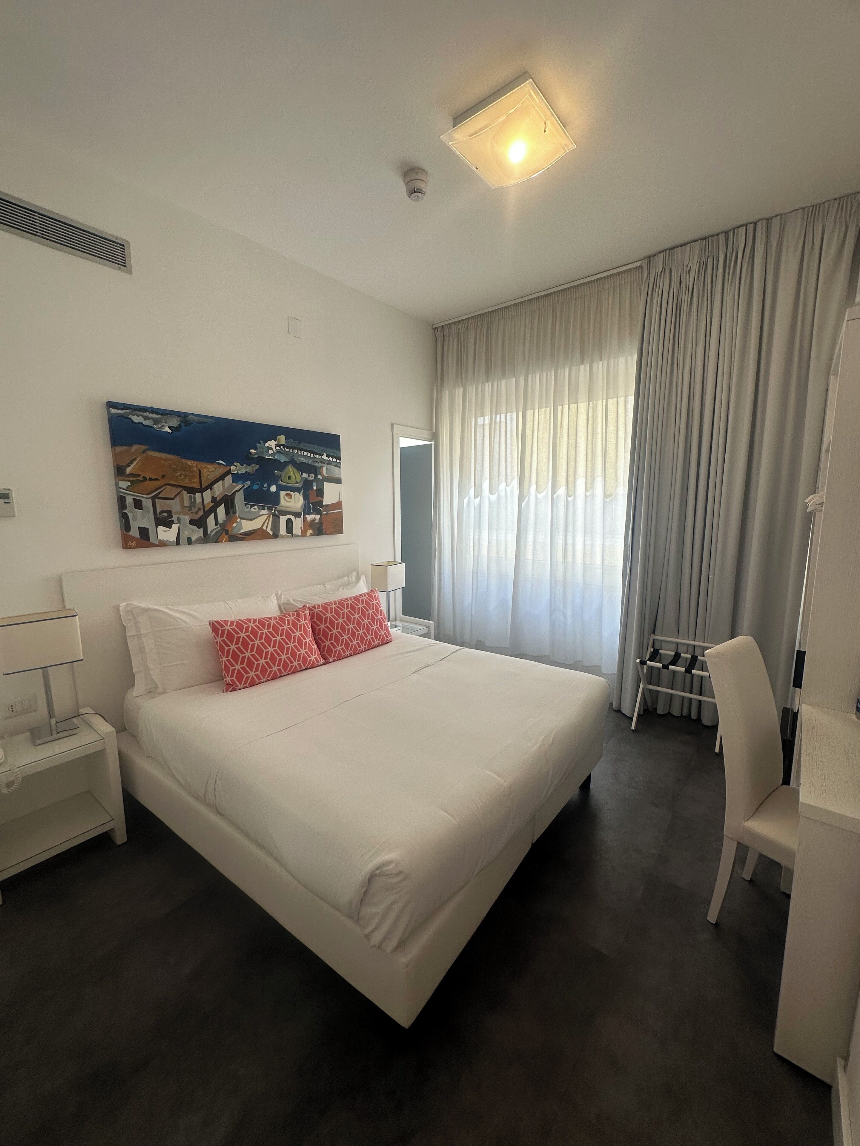 Habitación de hotel con una cama grande, una mesita de noche, una silla, un cuadro en la pared y una ventana con cortinas.