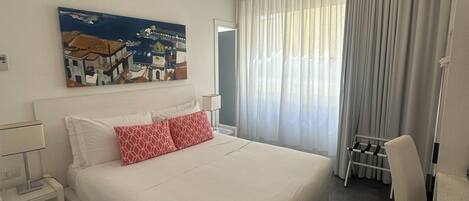 Habitación de hotel con una cama grande, una mesita de noche, una silla, un cuadro en la pared y una ventana con cortinas.
