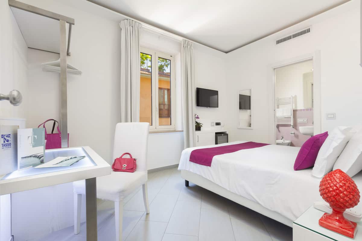 Habitación de hotel con cama, escritorio, silla, televisor, ventana con cortinas y baño visible a través de una puerta abierta.