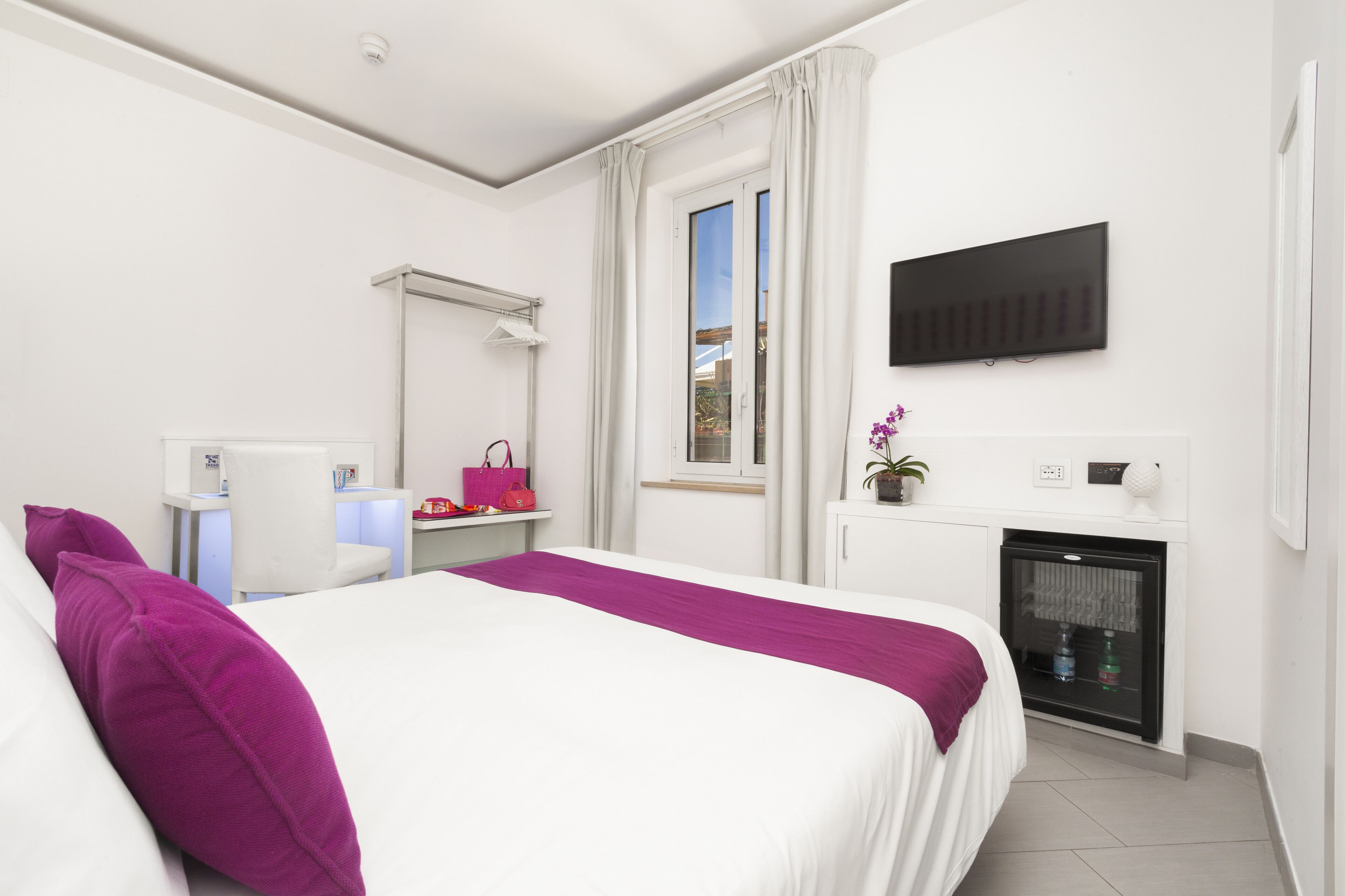 Une chambre d’hôtel équipée d’un lit, d’un bureau, d’une télévision, d’un mini-réfrigérateur et d’une fenêtre avec des rideaux.