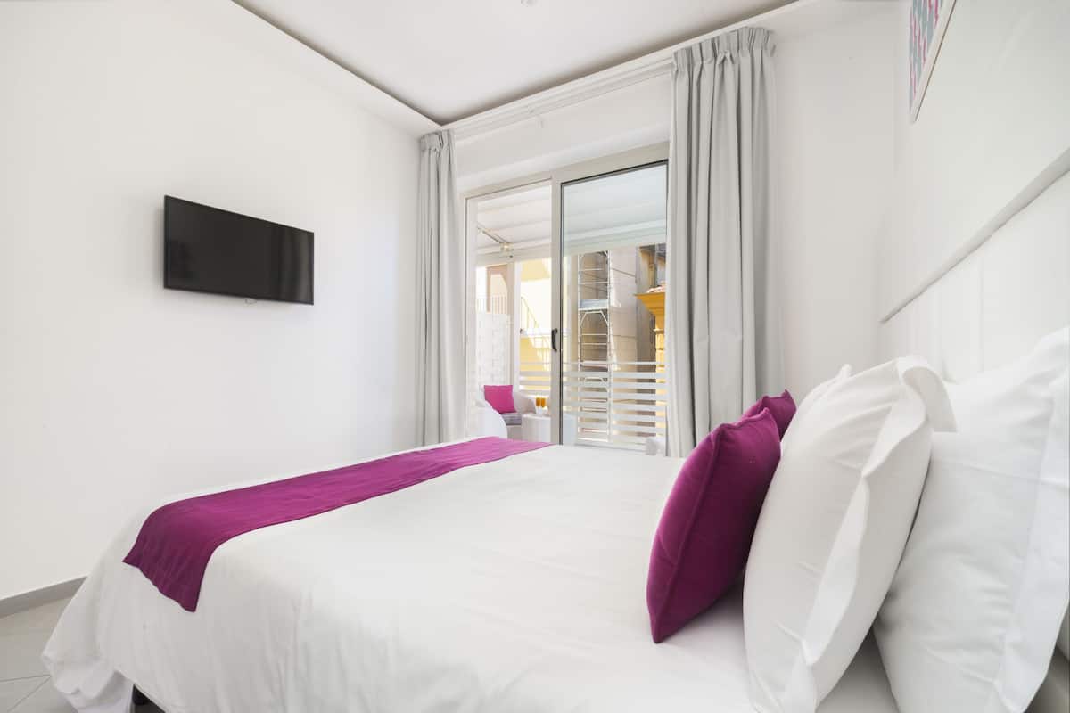 Habitación de hotel con cama, televisor y vista a un balcón con barandilla.