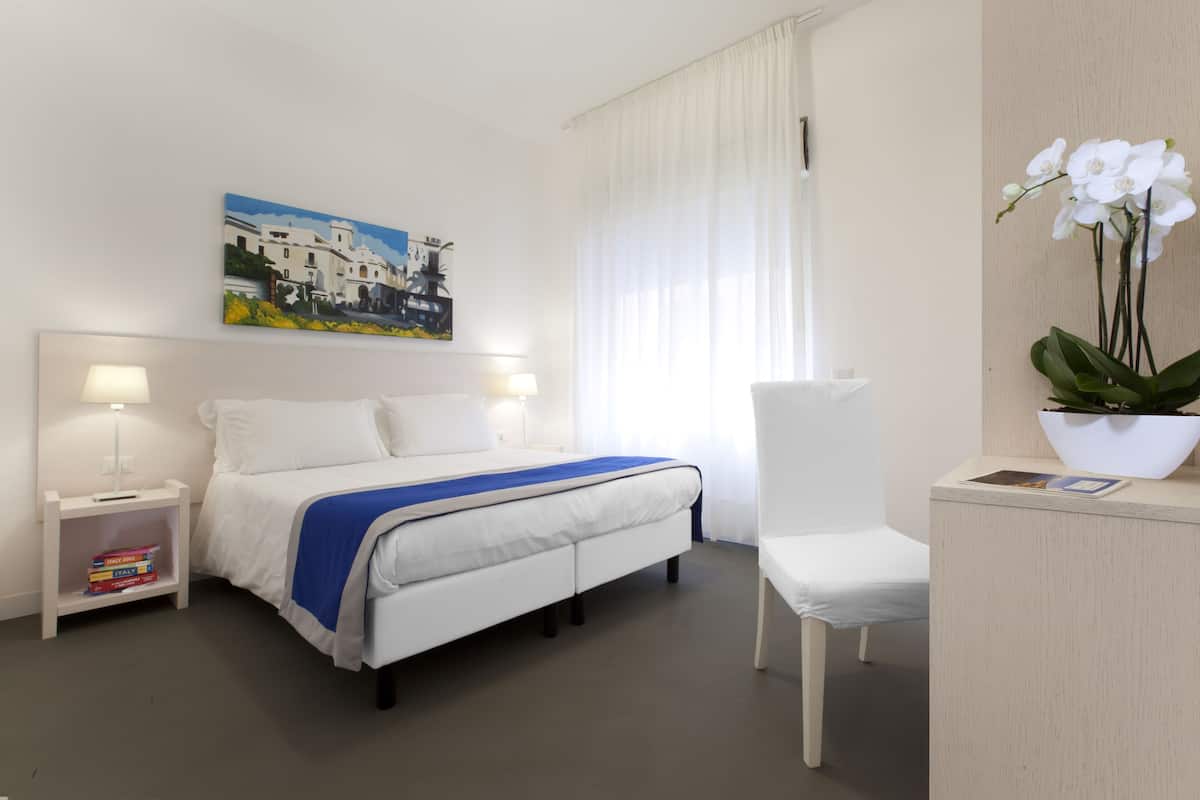 Habitación de hotel con cama, mesitas de noche, una silla, un escritorio, un cuadro y una planta en maceta.