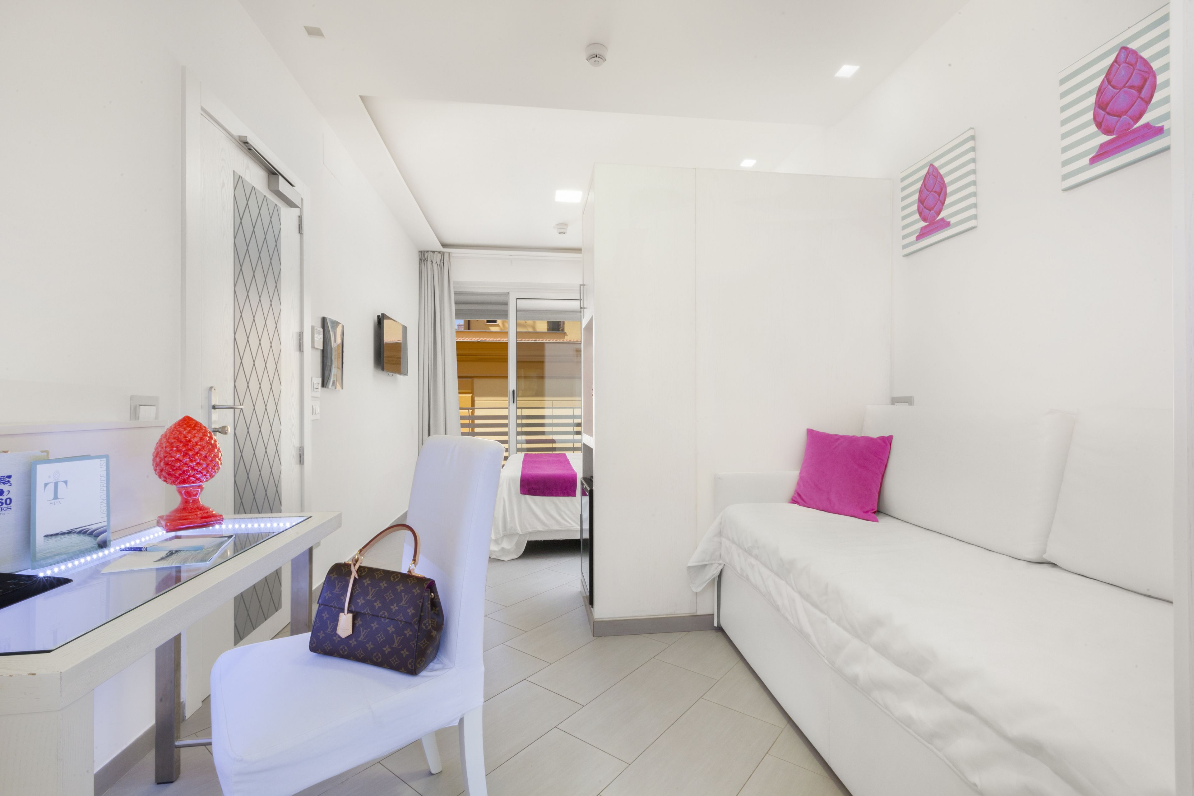 Quarto de hotel moderno com cama branca, uma poltrona branca, escrivaninha com computador e um objeto decorativo vermelho.