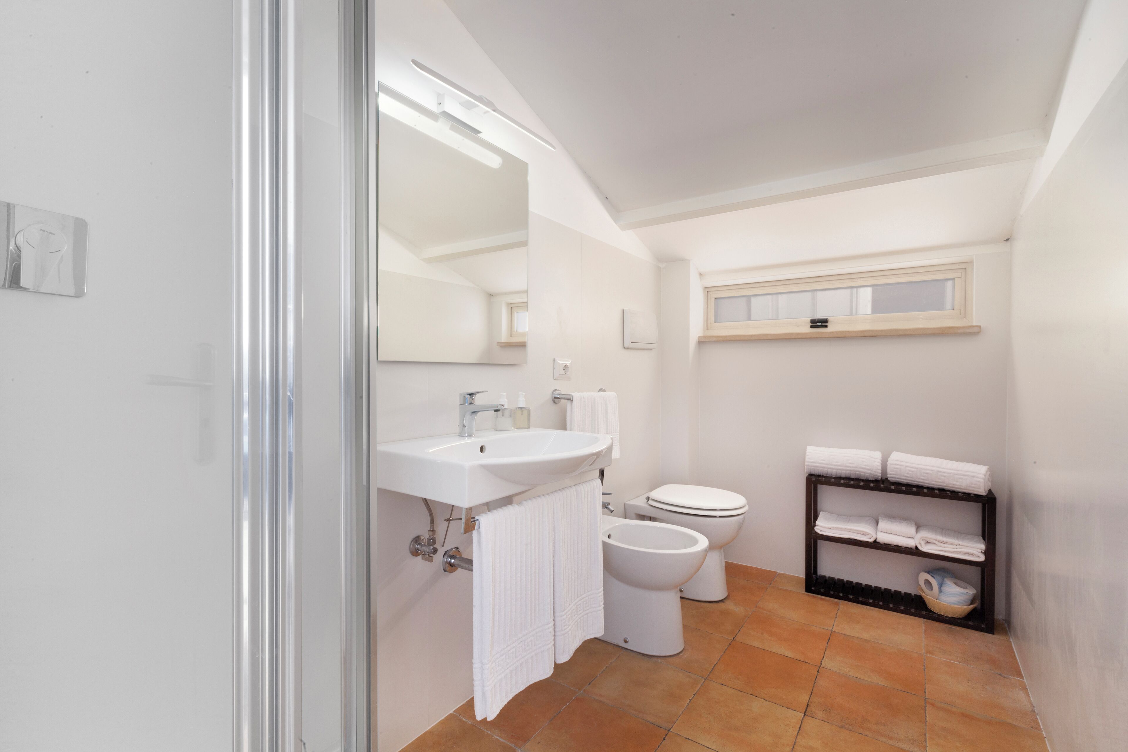 A modern bathroom with a sink, toilet, bidet, and a shelf with towels.