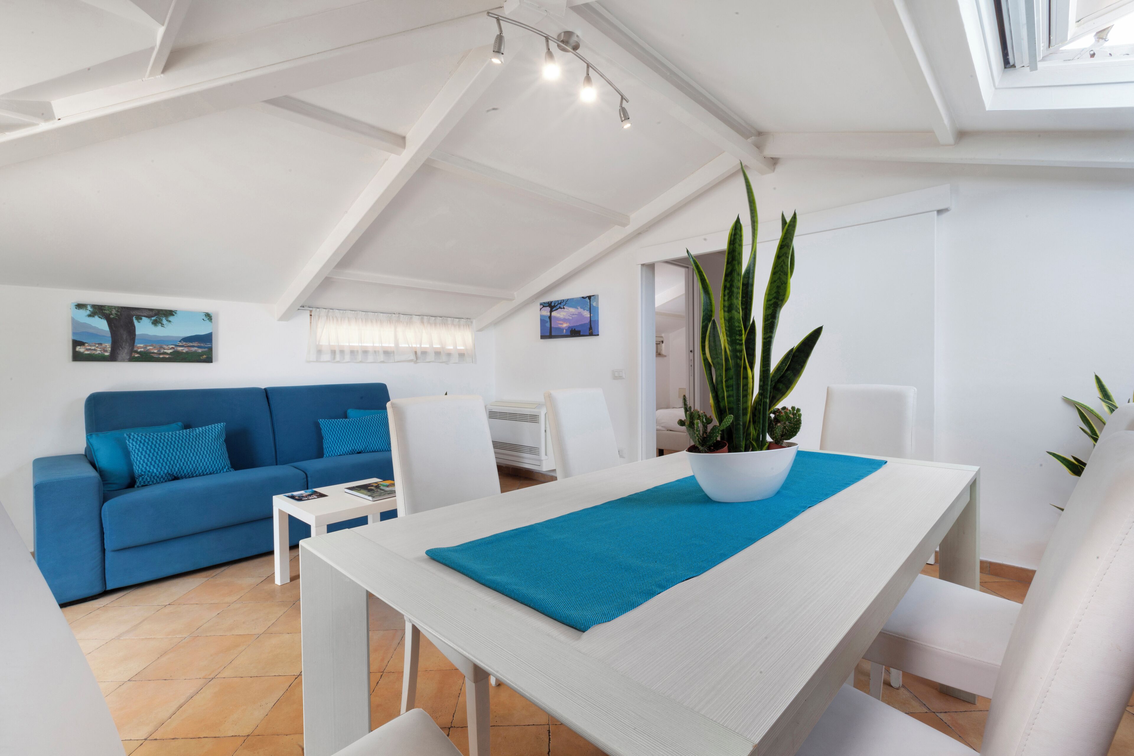 A modern living room with a blue sofa, white dining table, and a large potted plant.
