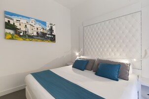 Un dormitorio con cama, almohadas, cabecero, mesita de noche y un cuadro en la pared.