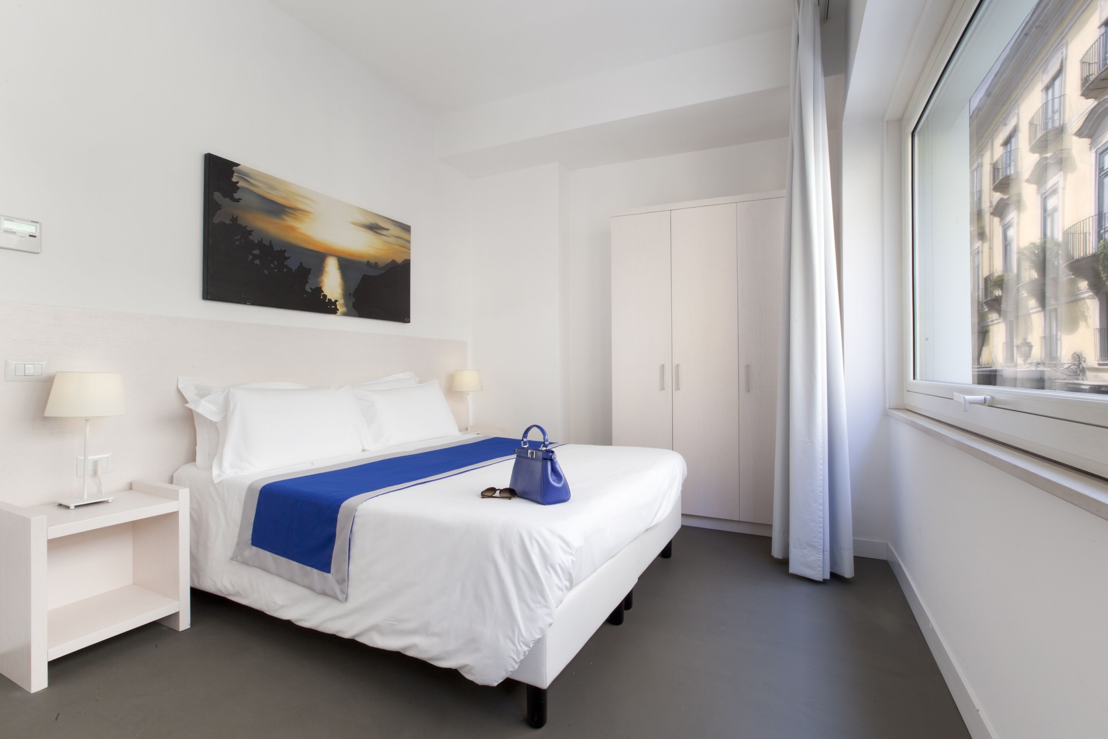 Habitación de hotel con cama, mesita de noche, lámpara, una bolsa azul sobre la cama, un cuadro en la pared y una ventana con vistas a edificios.