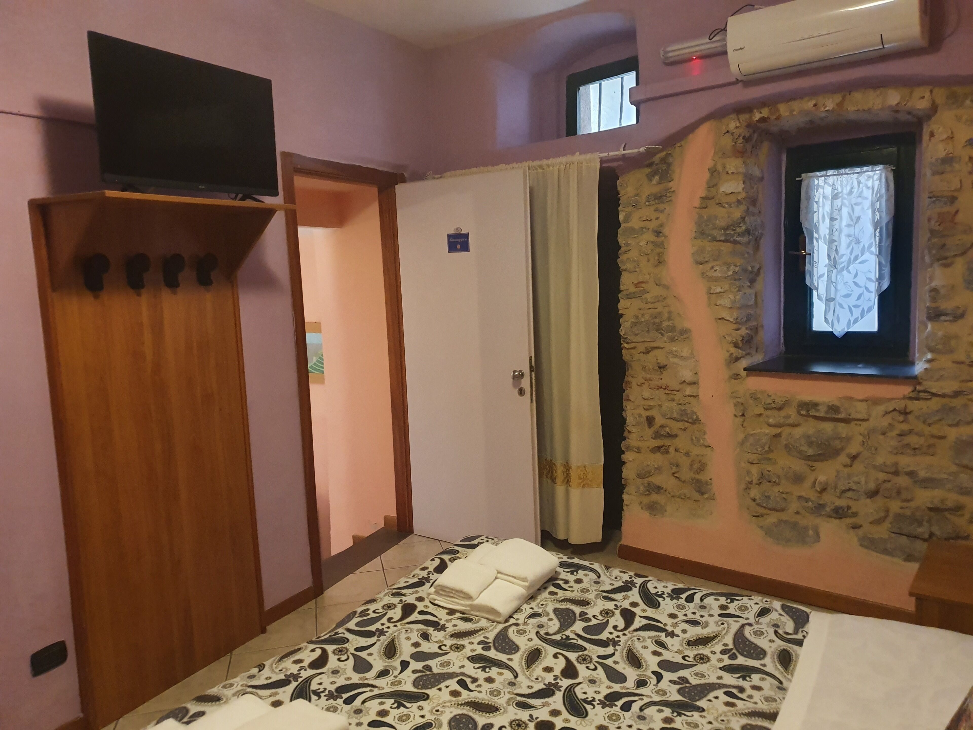 Une chambre à coucher avec un lit, une armoire en bois, un téléviseur posé sur un meuble, un mur en pierre et une fenêtre avec un rideau.