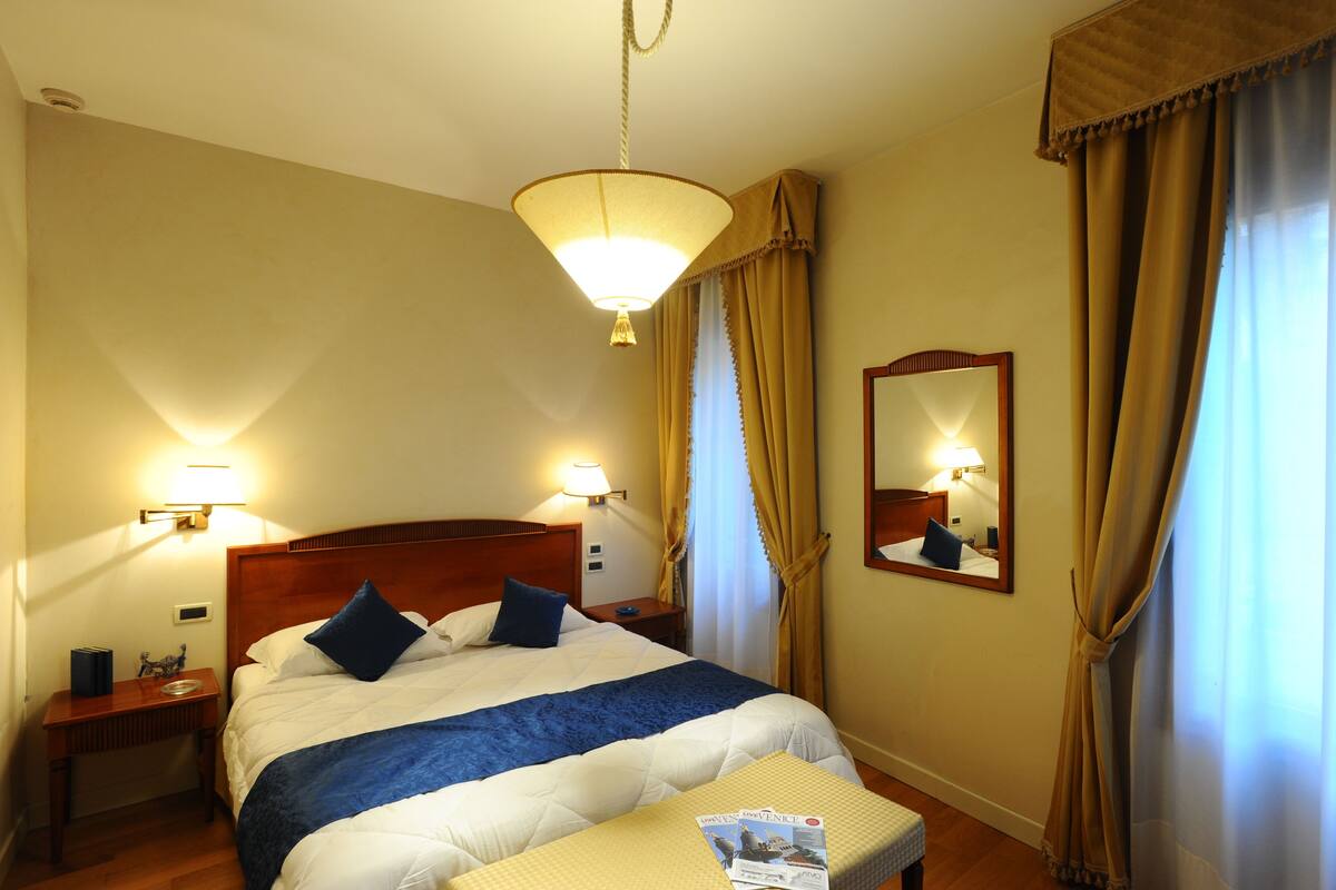 A hotel room with a large bed, two bedside lamps, a wooden nightstand, a small table with a magazine, and a mirror on the wall.