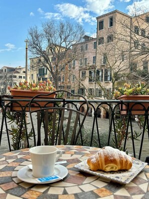 Una mesa con una taza y un pastel, suelo de baldosas y edificios al fondo.