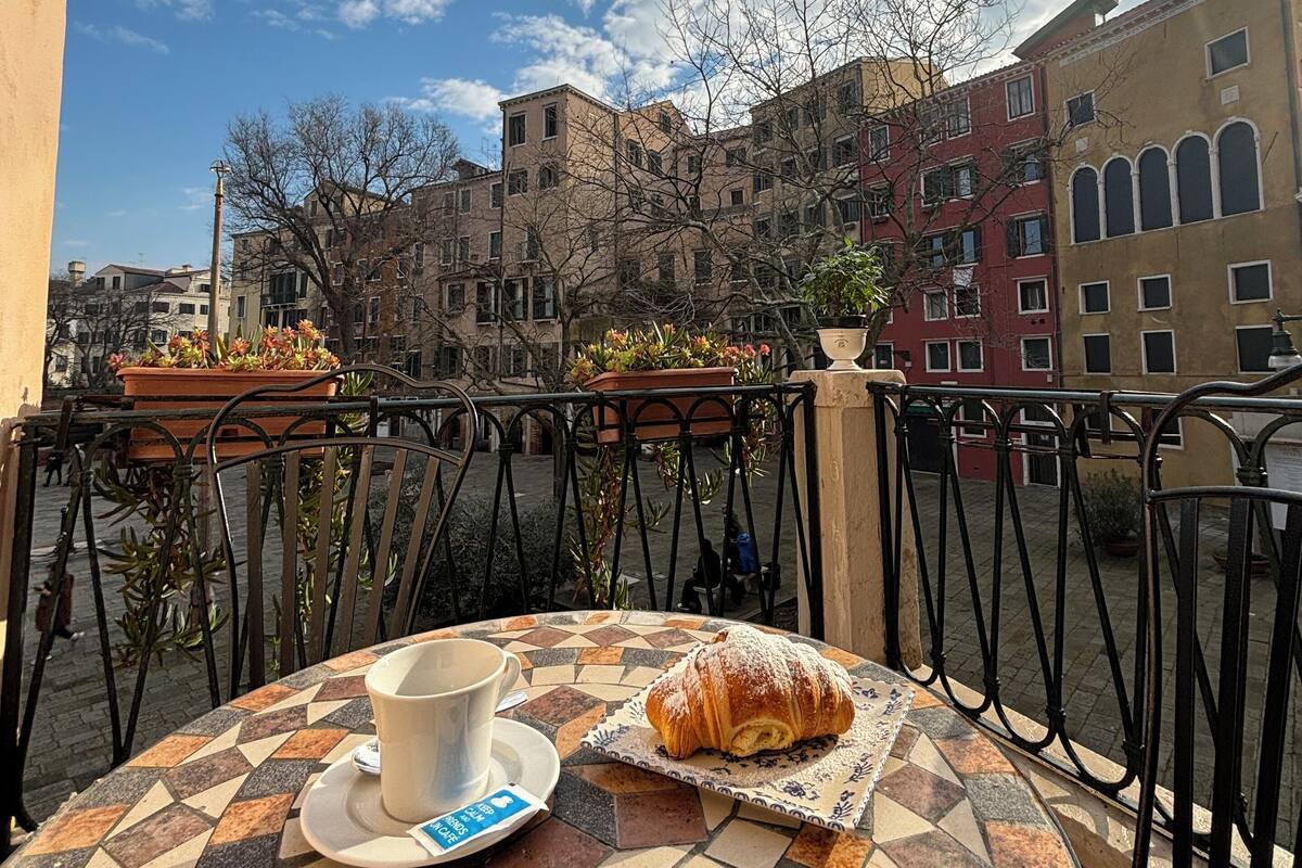 A round table with a croissant and a cup, overlooking a courtyard with buildings and a clear sky.