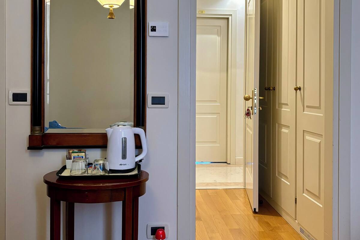 A hotel room with a wooden console table, a coffee maker, and a mirror. There is a door leading to a hallway with wooden flooring and a door with a number plate.
