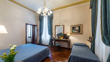 A hotel room with two beds, a desk, a chair, a chandelier, and a window with curtains.
