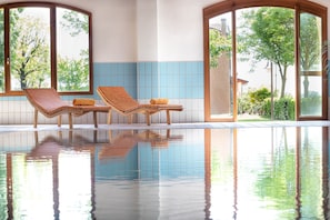 Une piscine avec deux chaises longues et une serviette, entourée de grandes fenêtres donnant sur un paysage verdoyant.