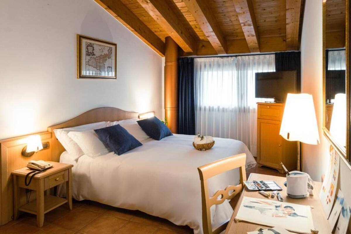 A hotel room with a wooden bed, a nightstand with a lamp, a desk with a lamp, a television, and a window with curtains.
