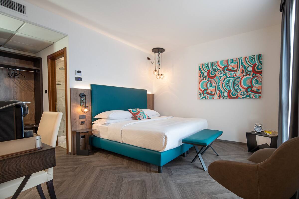 A modern hotel room with a teal bed, a wooden desk, a small table, a bench, and a wall-mounted artwork.