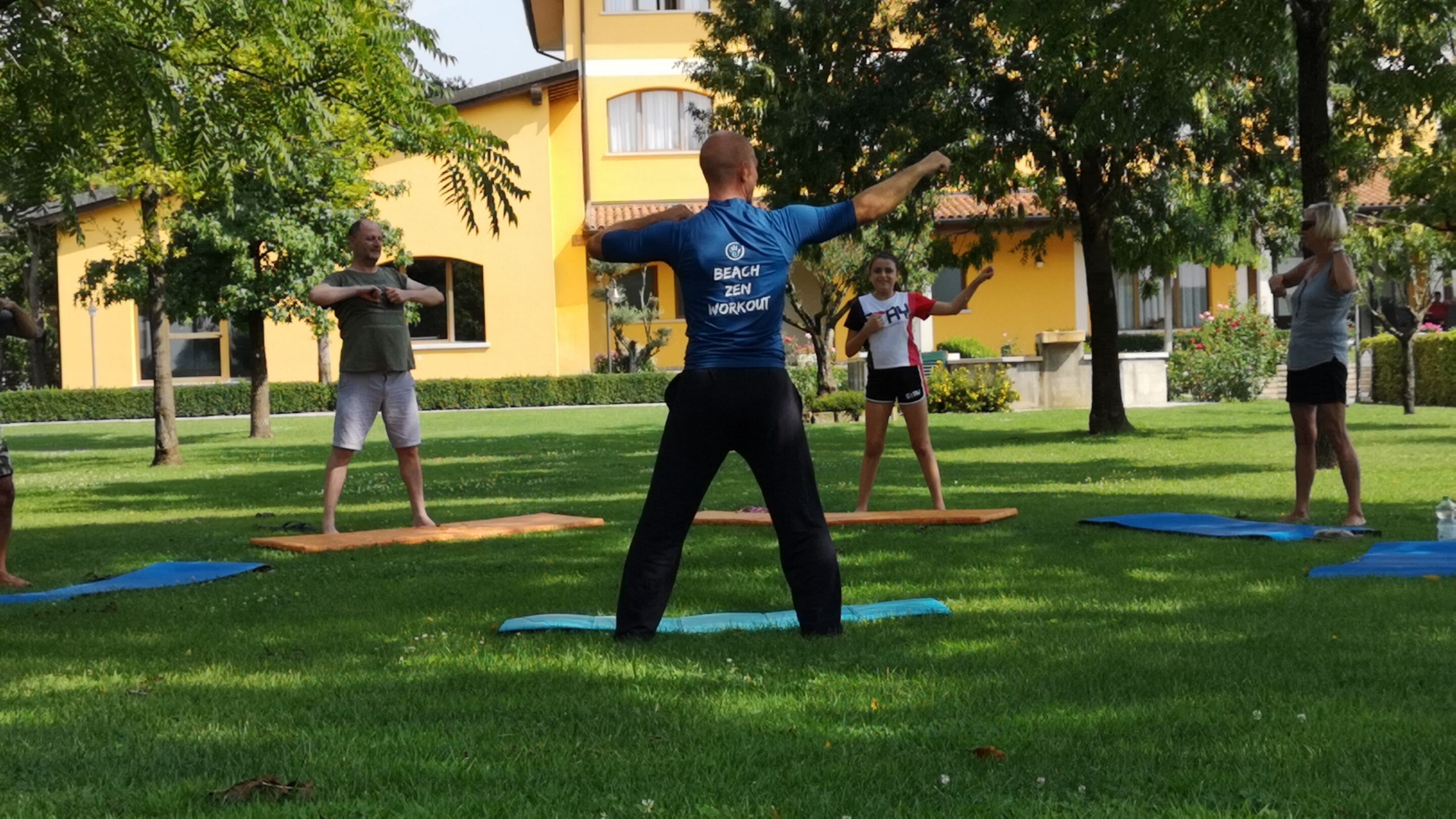 A group of people exercising outdoors on a grassy area with trees and a building in the background.