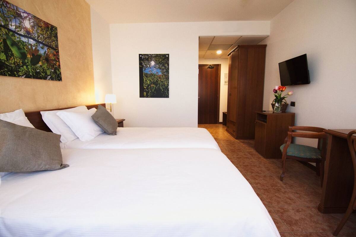 A hotel room with two beds, a desk, a chair, a TV, and artwork on the walls.