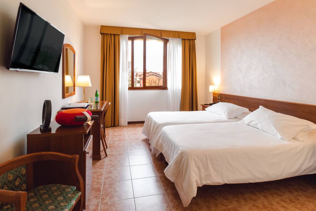 A hotel room with two beds, a desk with a chair, a TV mounted on the wall, and a window with curtains.