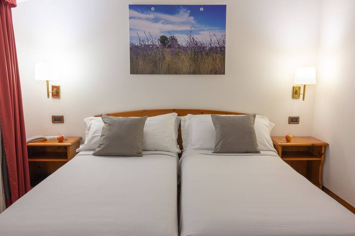 A double bed with two pillows, a nightstand with an apple, and a wall-mounted picture of a field.