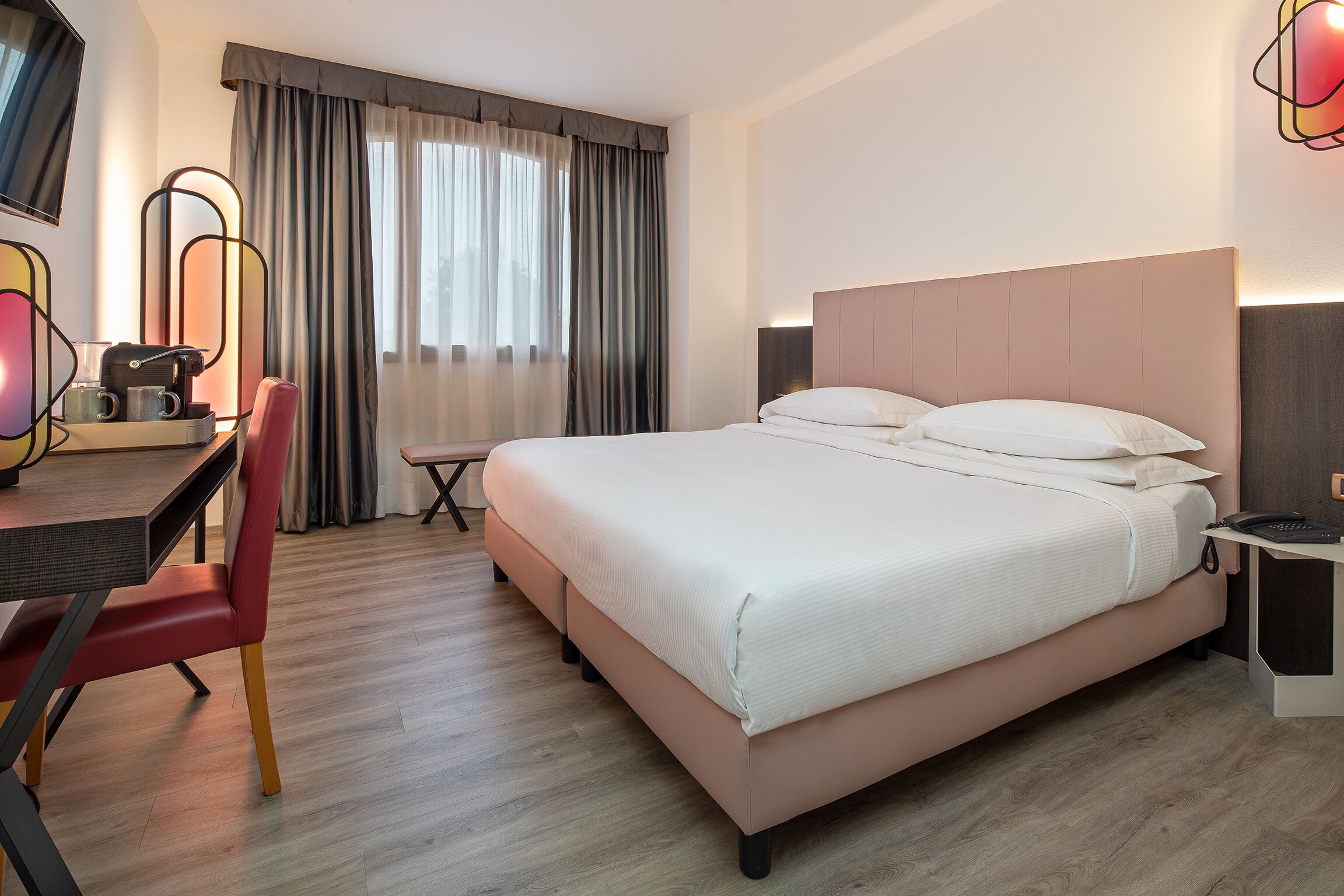 A hotel room with a bed, a desk with a chair, a TV, and a window with curtains.