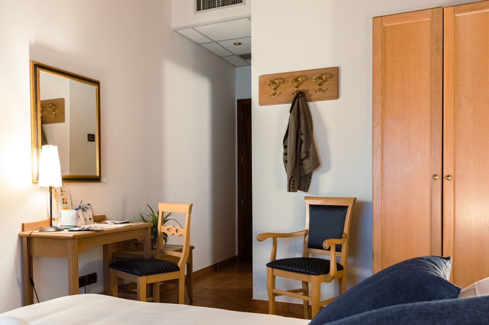 Et hotelværelse med seng, skrivebord med lampe, to stole, knagerække og garderobeskab i træ.
