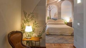Ein Zimmer mit Holzboden, einem Sessel, einer Lampe, einem kleinen Tisch und einem Bett mit Blick durch ein rundbogenförmiges Fenster.